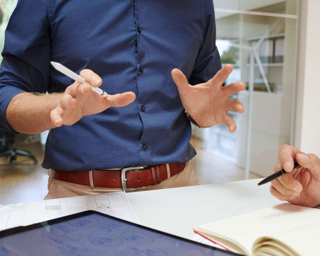 Zwei Projektingenieure tauschen sich in einem Büro aus und gestikulieren dabei mit Stiften in der Hand neben einem Tisch, auf dem sich ein Tablet und ein Notizbuch befinden