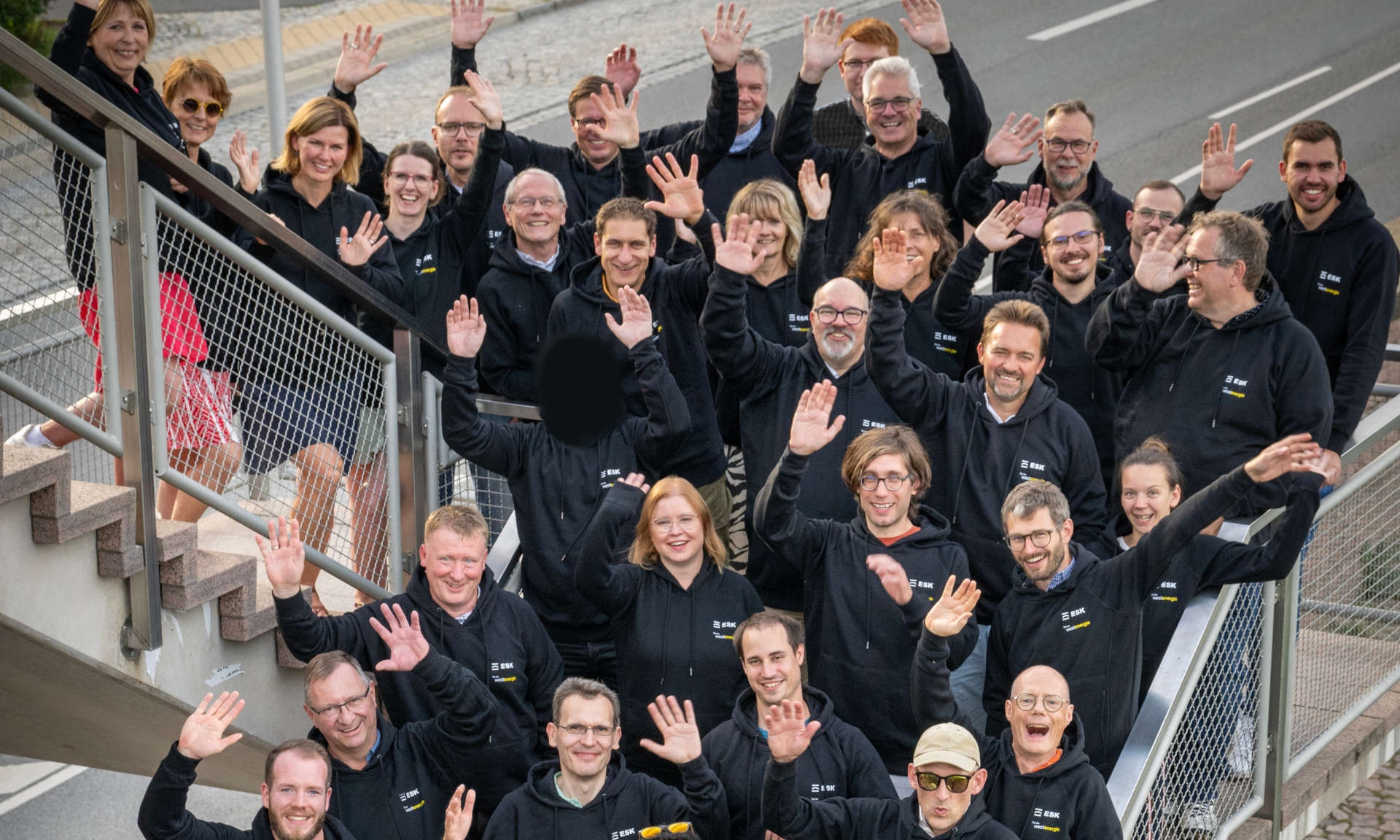 Winkendes Team der ESK GmbH in schwarzen Pullovern auf einer Treppe