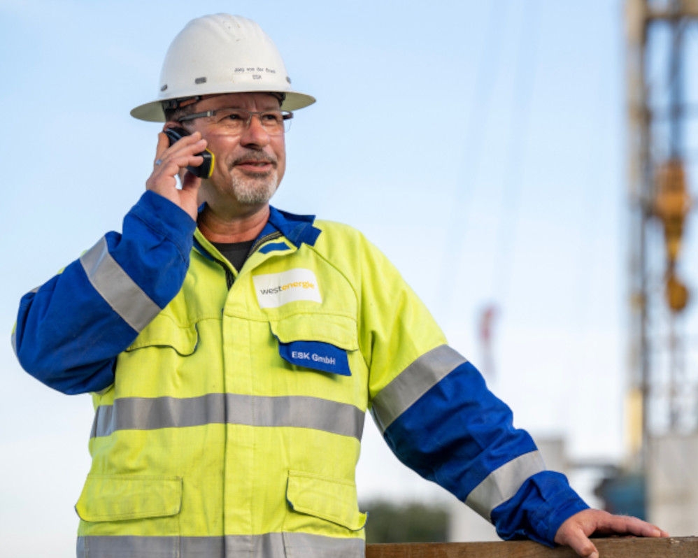 Ein Mann in Schutzkleidung und mit weißem Schutzhelm telefoniert auf der Baustelle 