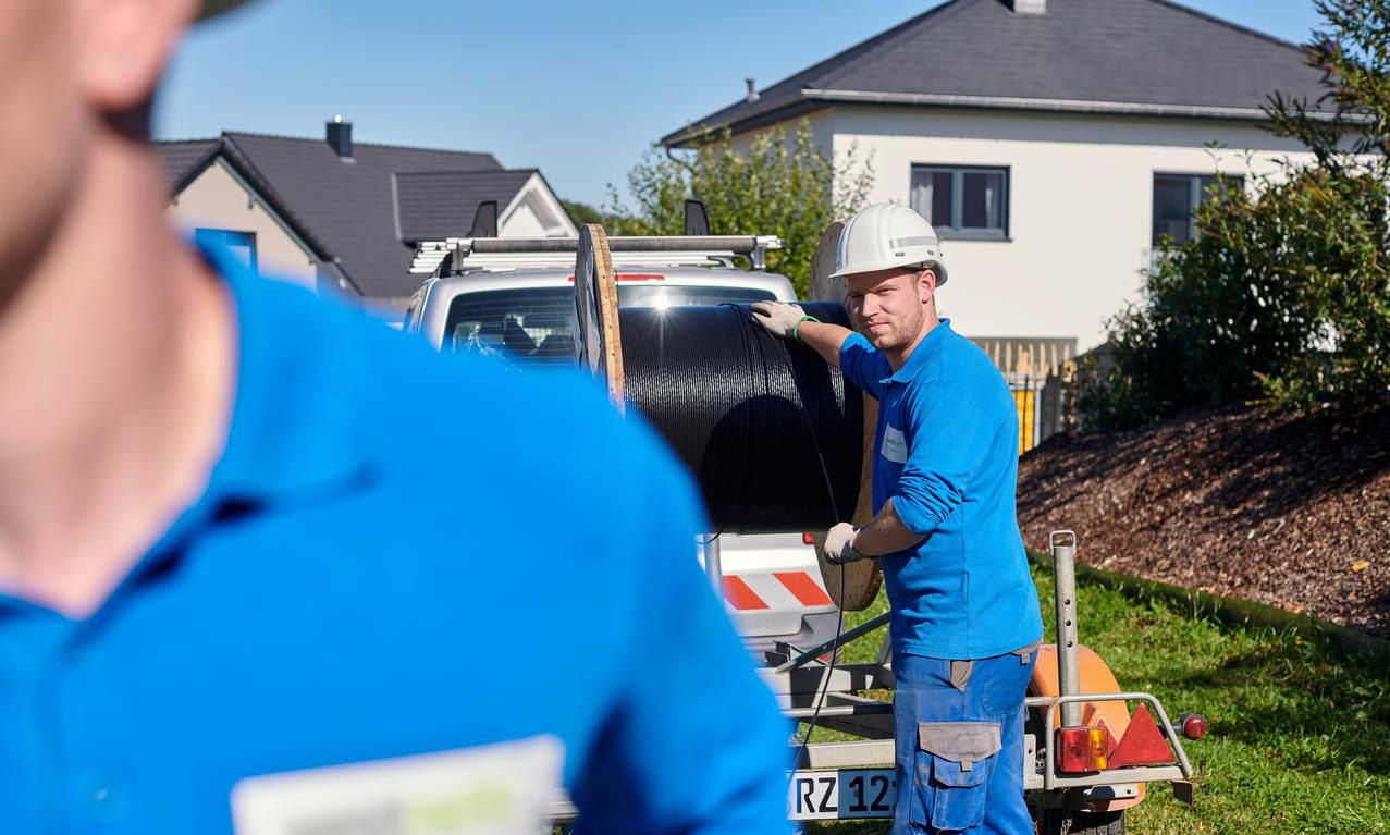 Employee in work clothes and a white safety helmet standing in a residential area behind a trailer with a cable reel on it
