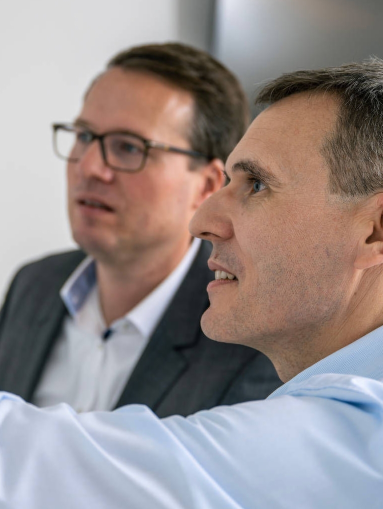 Two men are in an office in front of a whiteboard, one explaining something with a raised arm