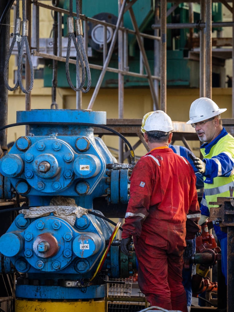 Three workers in protective clothing and white helmets having a conversation next to a blue wellhead