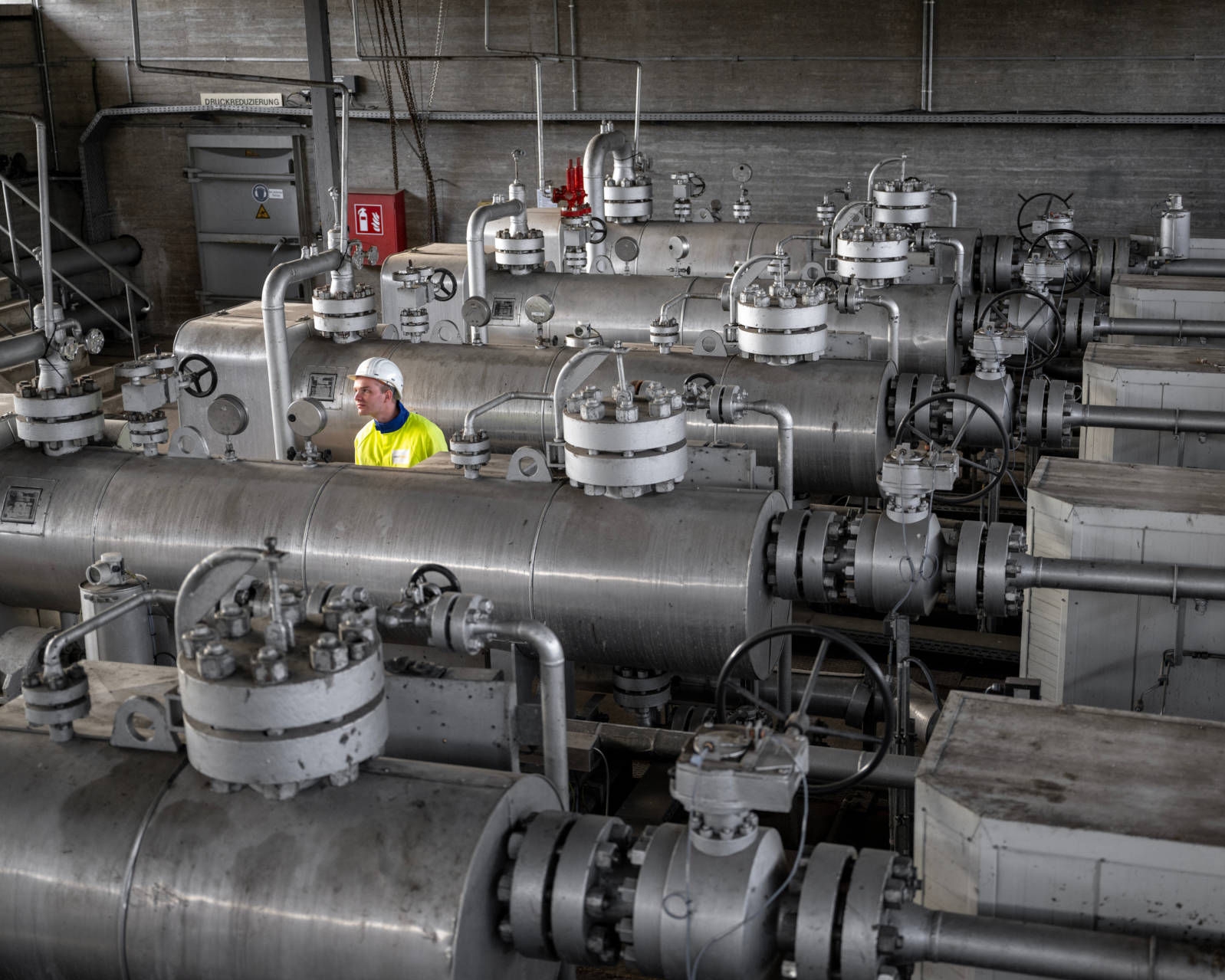 Working student in protective clothing and a white helmet at the methanol facility of an underground gas storage site, with large pipelines and valves 