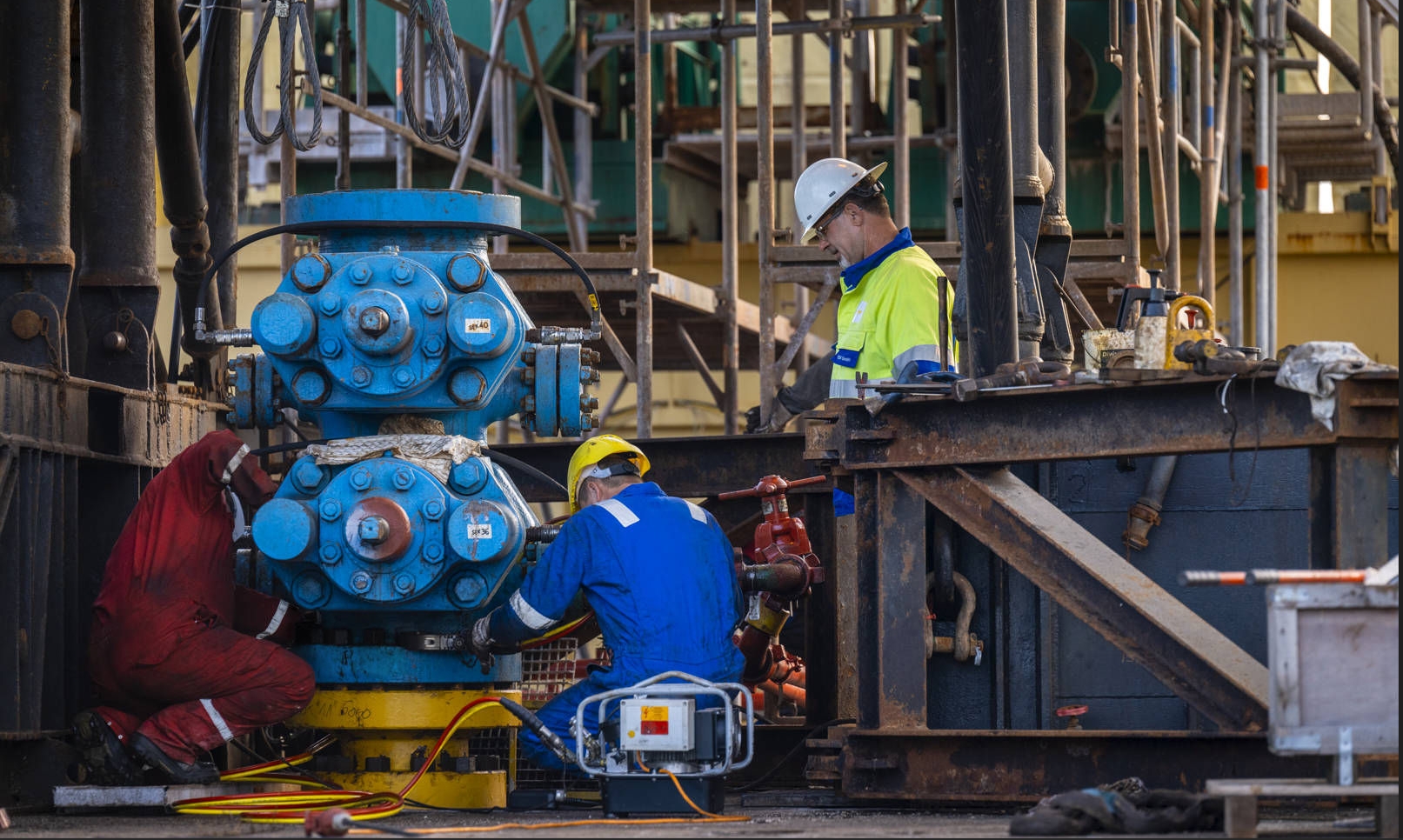 Three workers in protective clothing working on a wellhead with scaffolding in the background