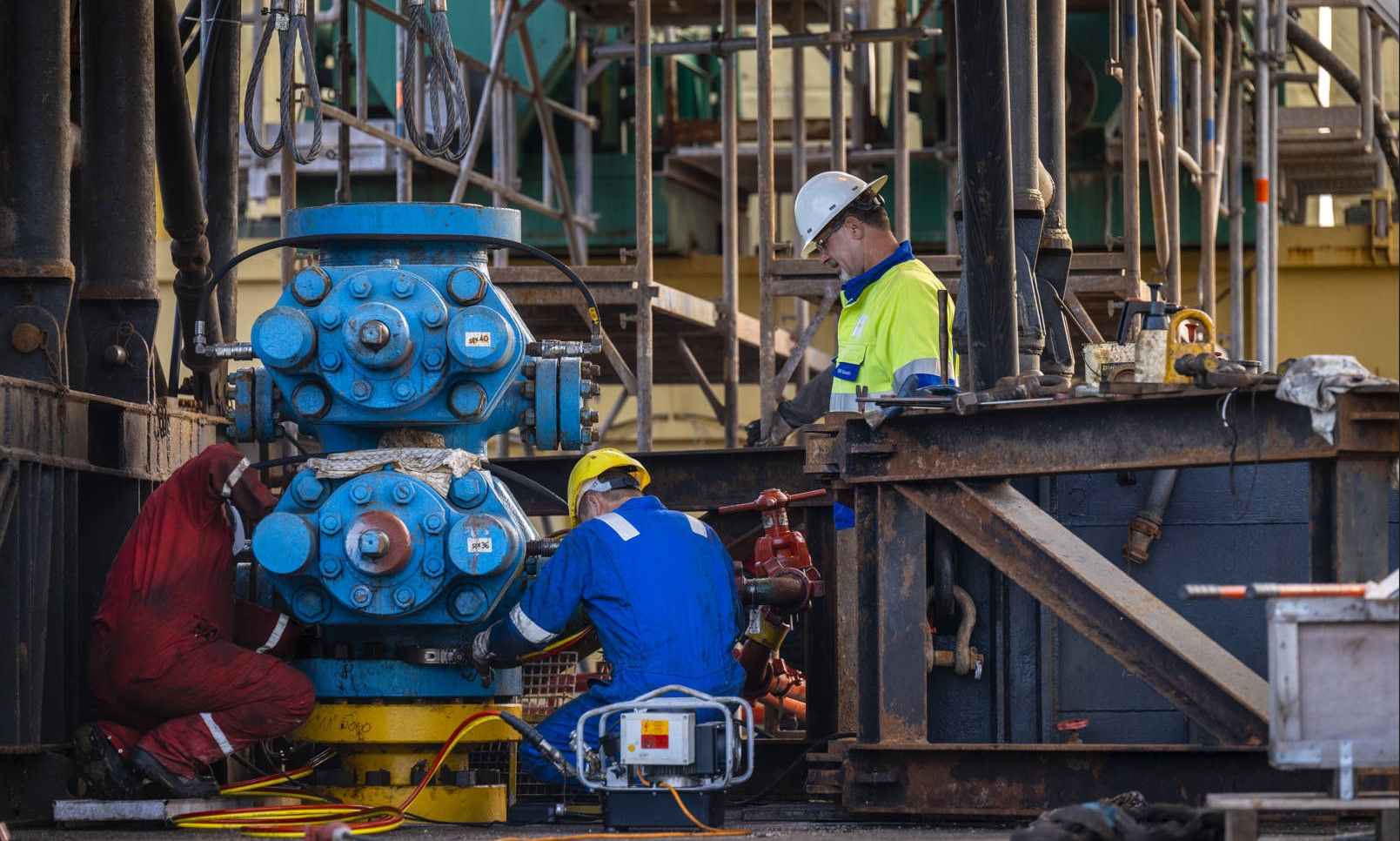 Three workers in protective clothing working on a wellhead with scaffolding in the background
