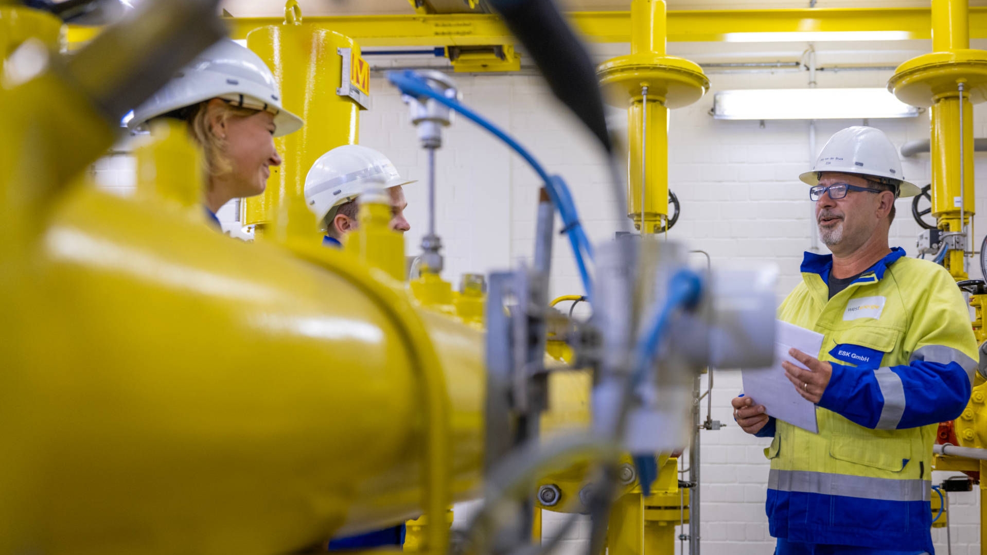 Three employees in protective clothing and white helmets are talking inside a gas storage facility (gas quantity measurement and analysis) with large yellow gas pipelines