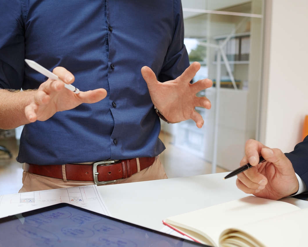Two project engineers are exchanging ideas in an office, gesturing with pens in hand, next to a table with a tablet and a notebook on it
