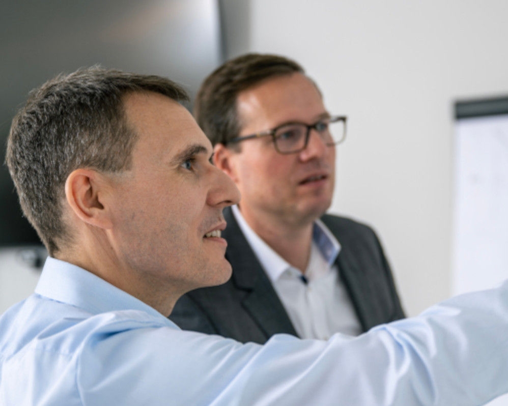 Two men in side profile are looking in the same direction in an office. The one in the foreground is pointing to a spot with his arm