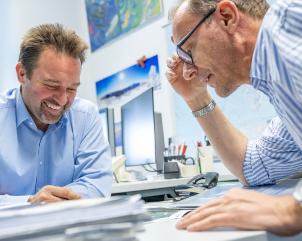 Two smiling project engineers in blue shirts are sitting across from each other at a desk with documents in an office. In the background are pens, screens, and pictures on the wall