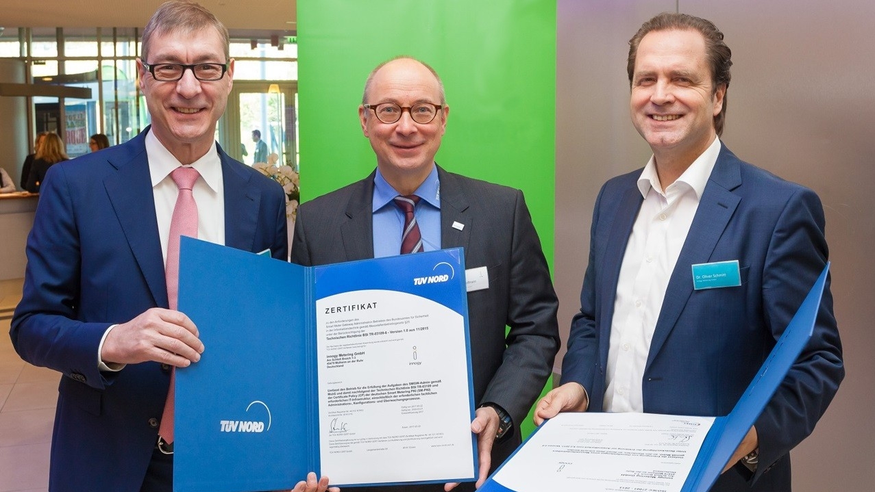 3 Männer in Business-Anzügen schauen in die Kamera und lächeln, sie halten ein TüV-Zertifikat in die Höhe